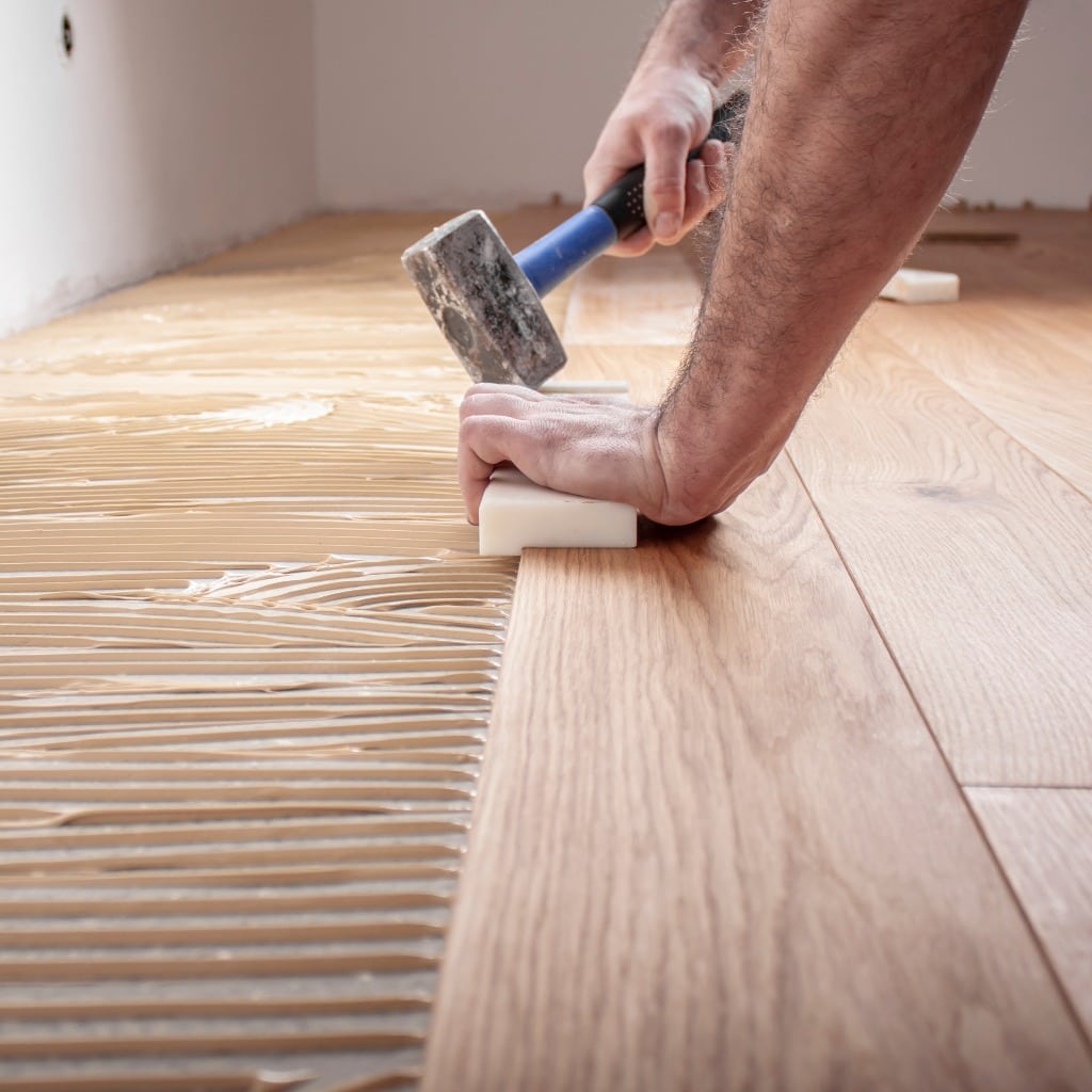 Handwerker verlegt Parkettboden und streicht den Estrich mit Kleber ein und schlägt die Dielen mit dem Hammer und dem Schlagklotz zusammen
